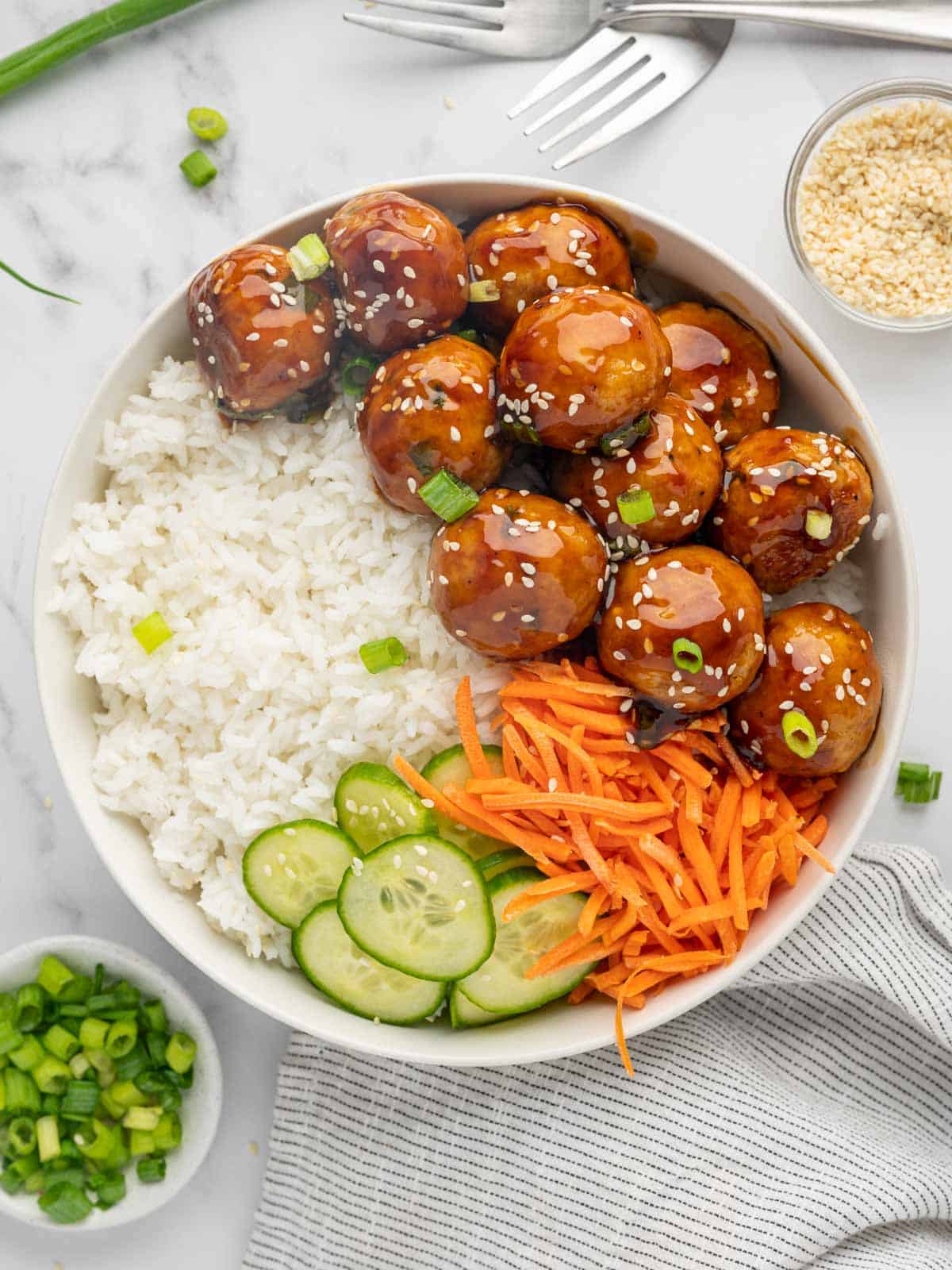 Teriyaki meatballs on a plate with rice, sliced cucumber and julienned carrots.