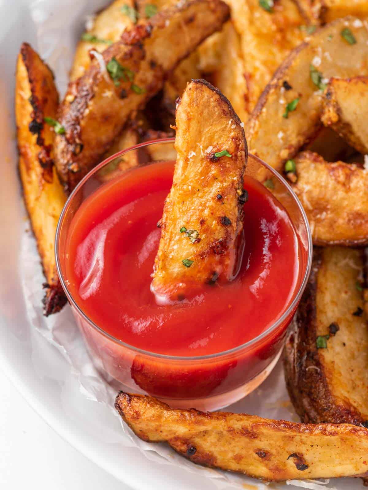 A plate of potato wedges with one dipping into ketchup.