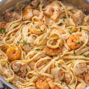 A skillet with pasta, chicken and shrimp topped with alfredo sauce.