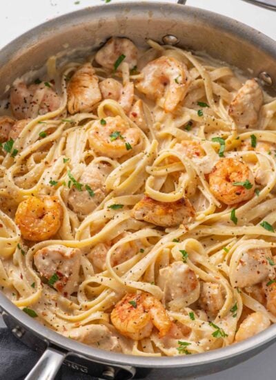A skillet with pasta, chicken and shrimp topped with alfredo sauce.