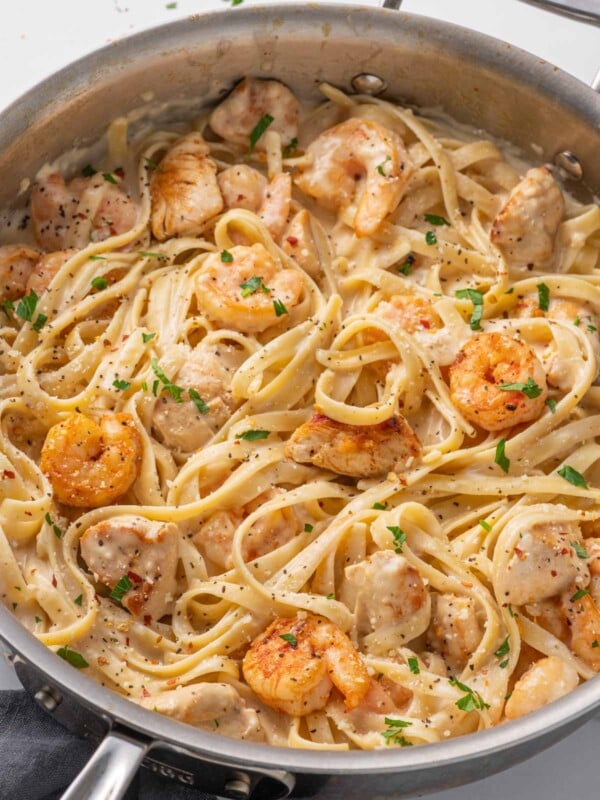 A skillet with pasta, chicken and shrimp topped with alfredo sauce.