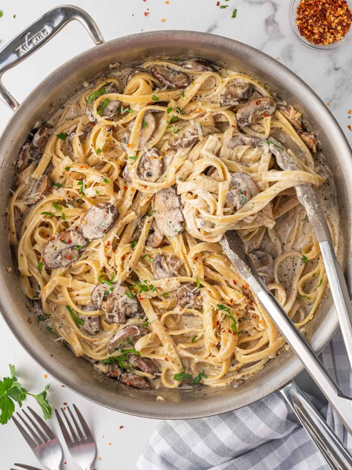 Mushroom pasta is being stirred in skillet with tongs.