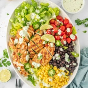 Ingredients for grilled chicken salad recipe piled on a plate with forks and dressing to the side.