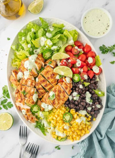 Ingredients for grilled chicken salad recipe piled on a plate with forks and dressing to the side.