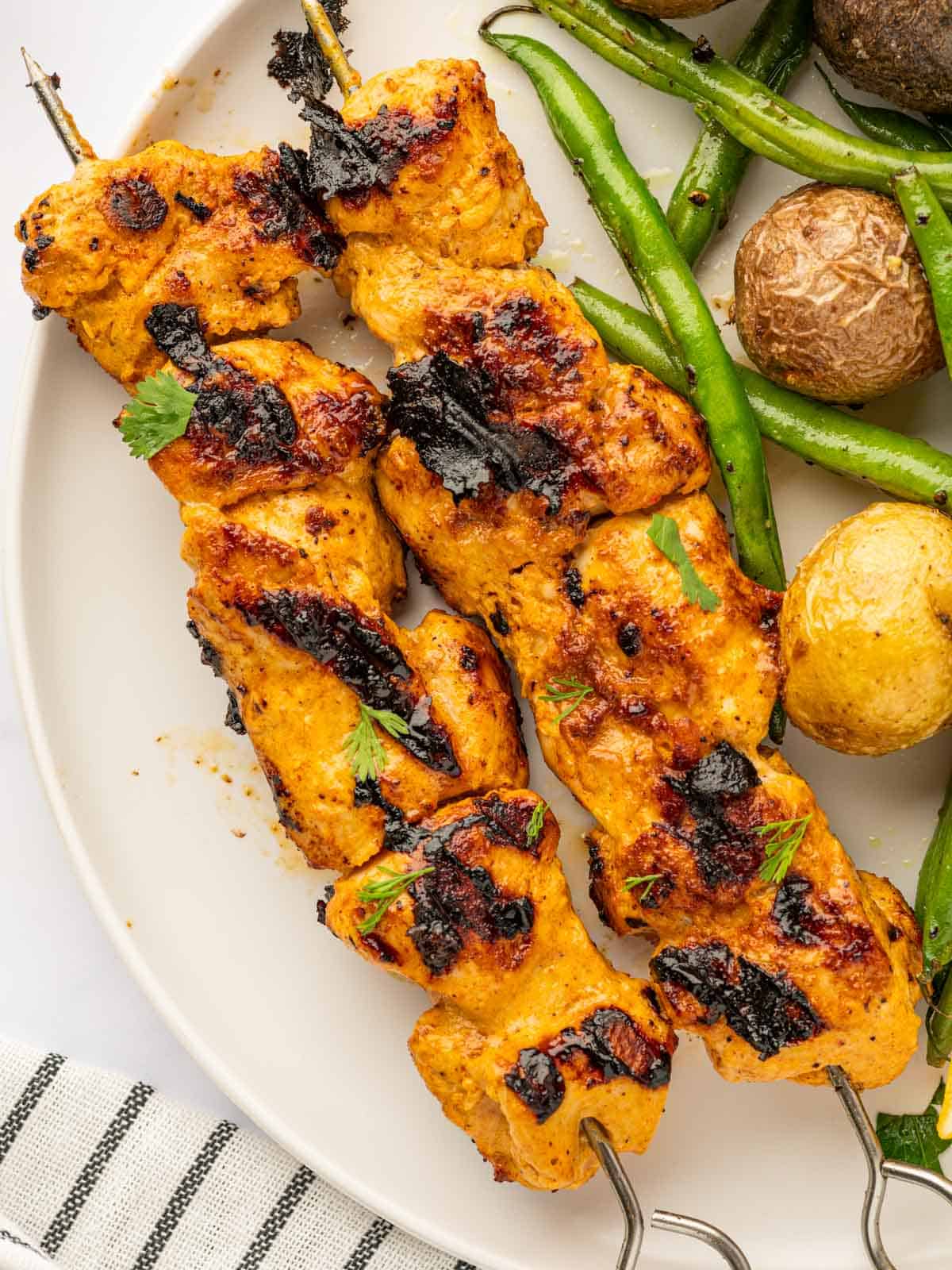 Tandoori chicken skewers on a plate with green beans and roasted potatoes.