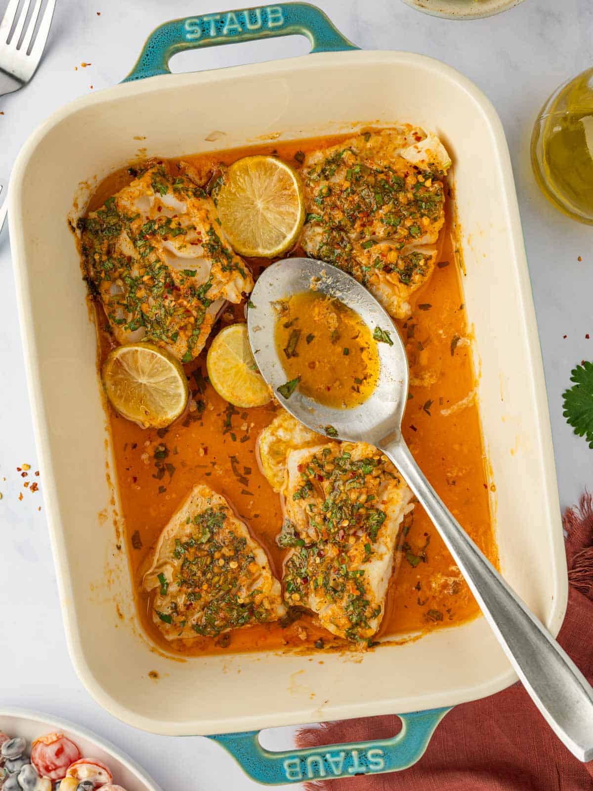 A spoon ladles buttery herb lime cod marinade over cooked fish in a casserole dish.