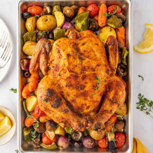Golden whole chicken surrounded by roasted potatoes, carrots, tomatoes, mushrooms, peppers, onions, and olives.