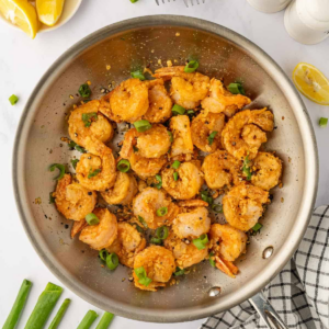 Overhead view of golden, crispy shrimp with garlic and sliced green onions in a stainless steel skillet.