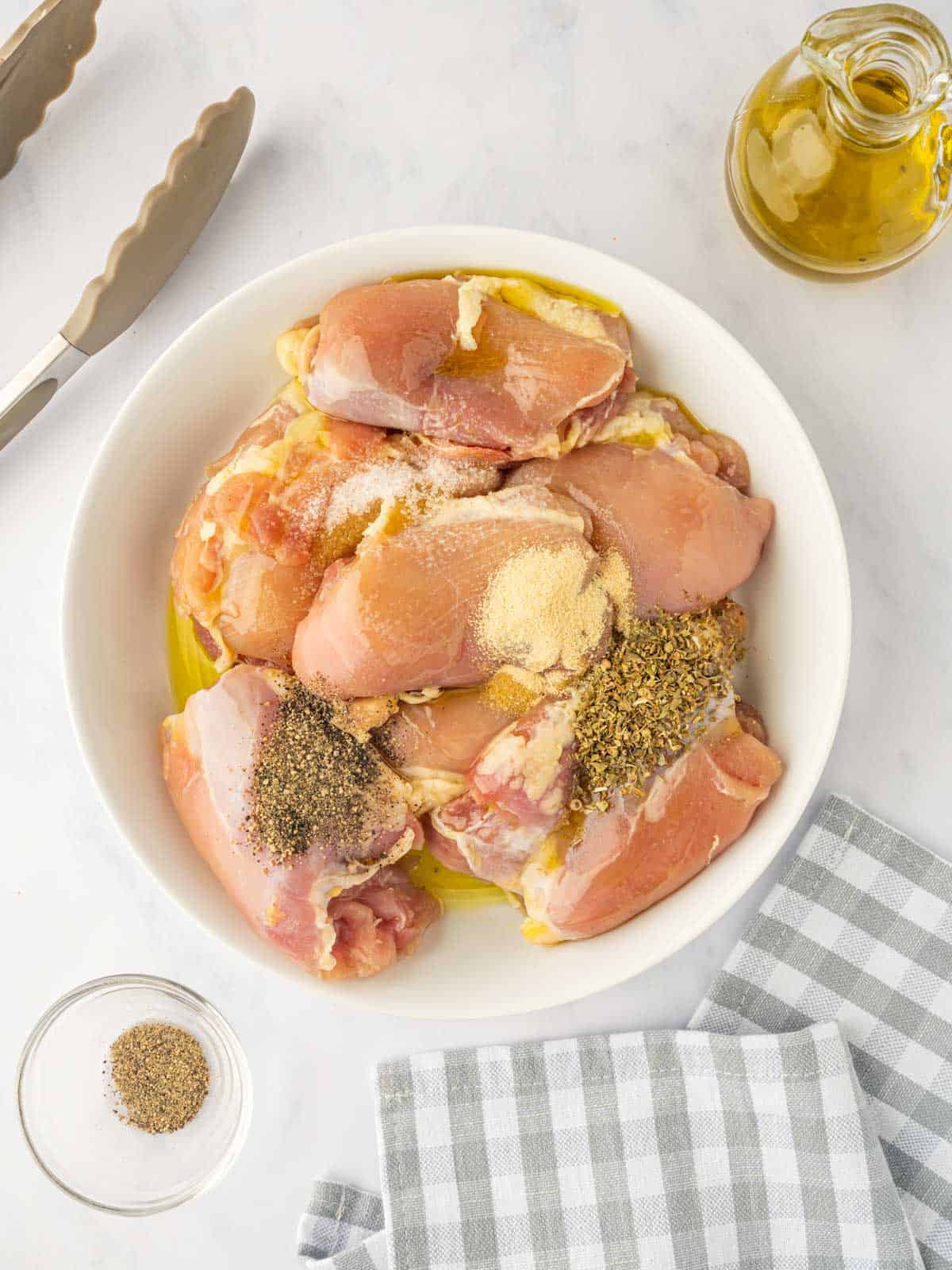 Raw chicken thighs are tossed with oil and seasoning in a bowl.