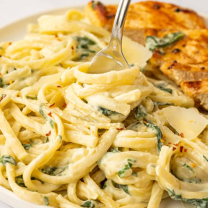Close-up of creamy linguine with spinach and Parmesan shavings, with a fork twirling the pasta and grilled chicken in the background.