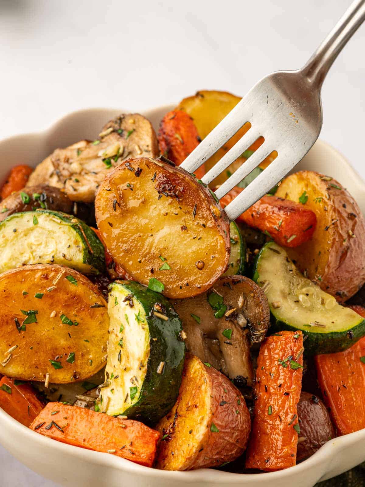 A fork picks up a roasted potato from a bowl of roasted vegetables.