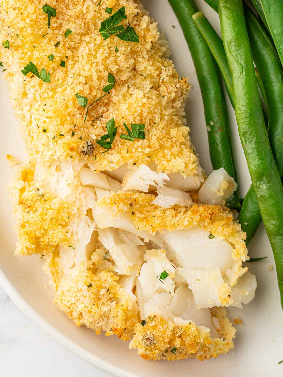 A piece of flaked panko baked cod on a plate with green beans.