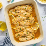 Baked chicken tenders in golden lemon sauce with lemon slices and chopped herbs.