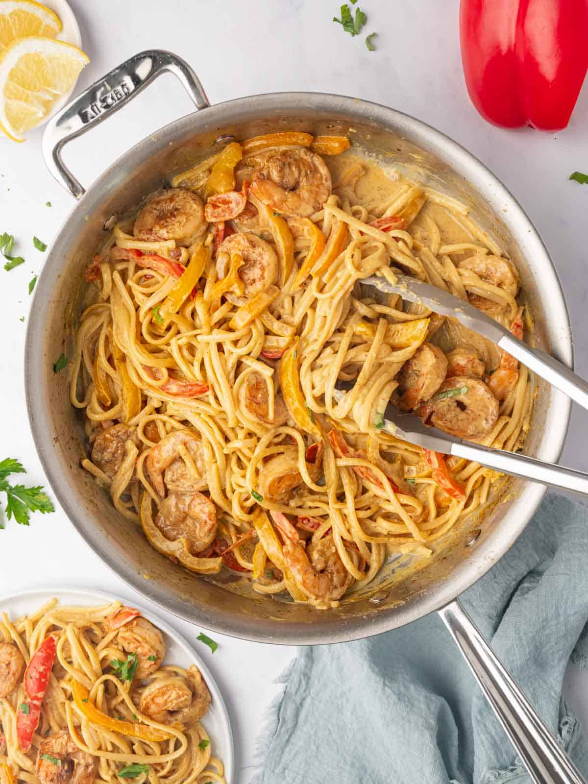 Tongs scoop servings of jamaican rasta pasta with shrimp from a skillet.