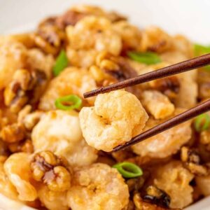 Chopsticks lifting a crispy fried shrimp over candied walnuts and sliced scallions.