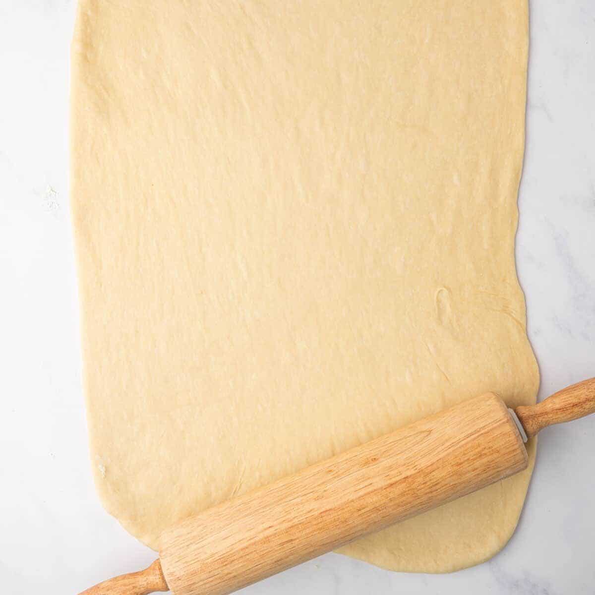 Cinnamon roll dough is rolled into a large rectangle with a wooden rolling pin.