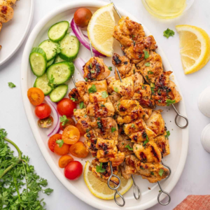 Grilled lemon pepper chicken kabobs on a platter with cucumbers, tomatoes, red onion, and lemon.