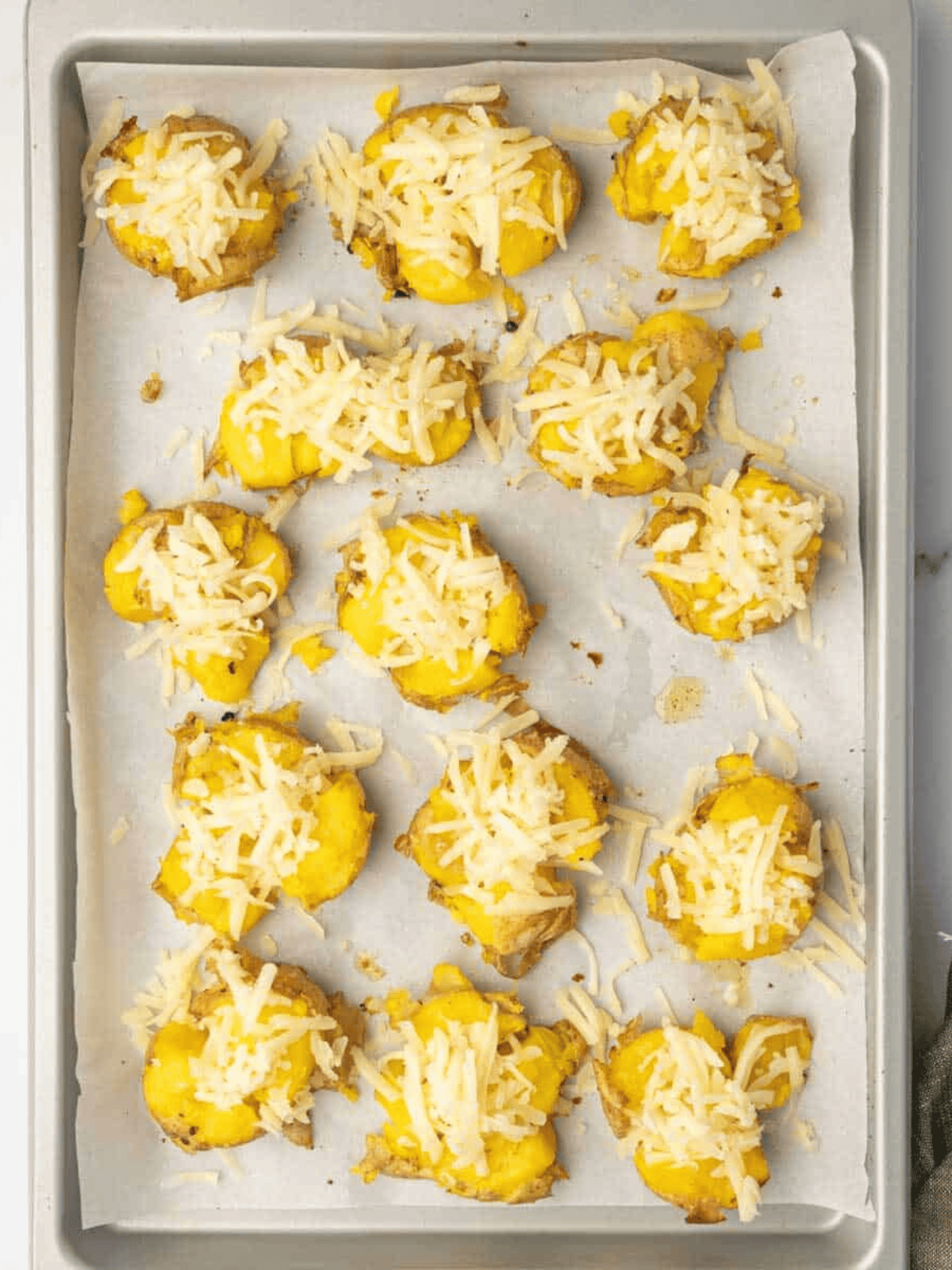 Smashed potatoes topped with shredded mozzarella on a parchment-lined baking tray before baking.