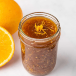 Homemade orange chicken sauce stored in a glass jar beside fresh oranges.