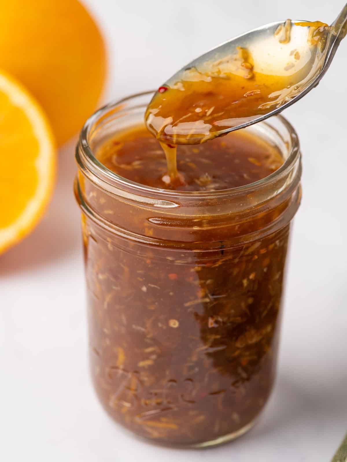 Orange chicken sauce dripping from a spoon over a glass jar.