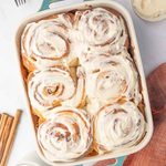 Cinnamon rolls covered with thick cream cheese icing in a white baking dish.
