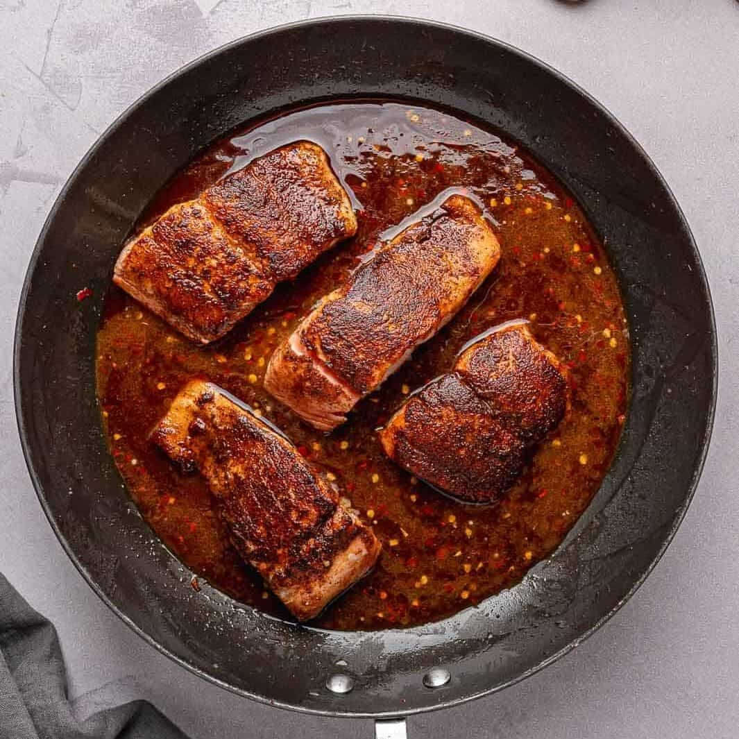 A dark, glossy honey garlic sauce over the seared salmon fillets in the pan, instantly bubbling as it hits the hot skillet.