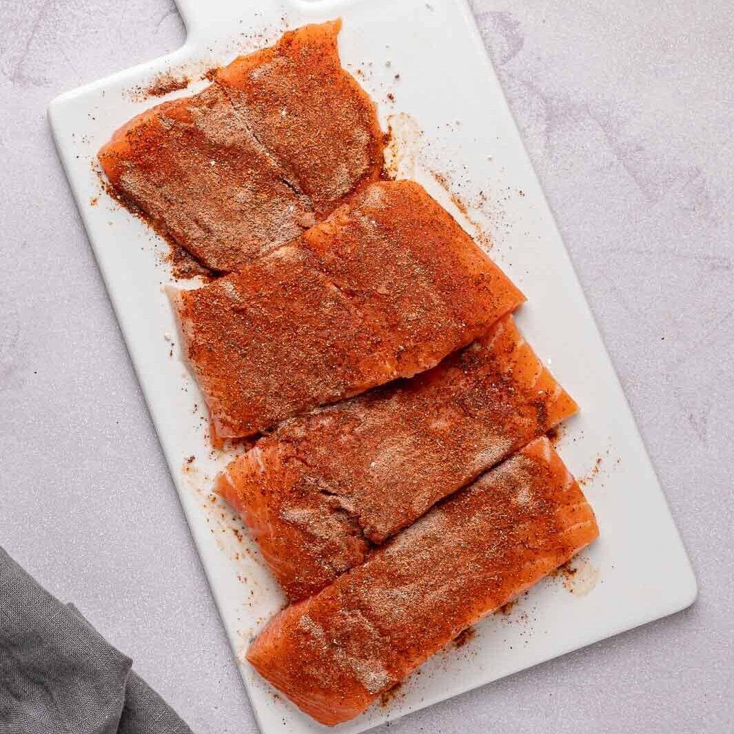 A close-up shot of four fresh salmon fillets laid out on a clean cutting board. Each one is completely covered in that vibrant, deep red spice rub, giving them a rich, dry, seasoned coating.