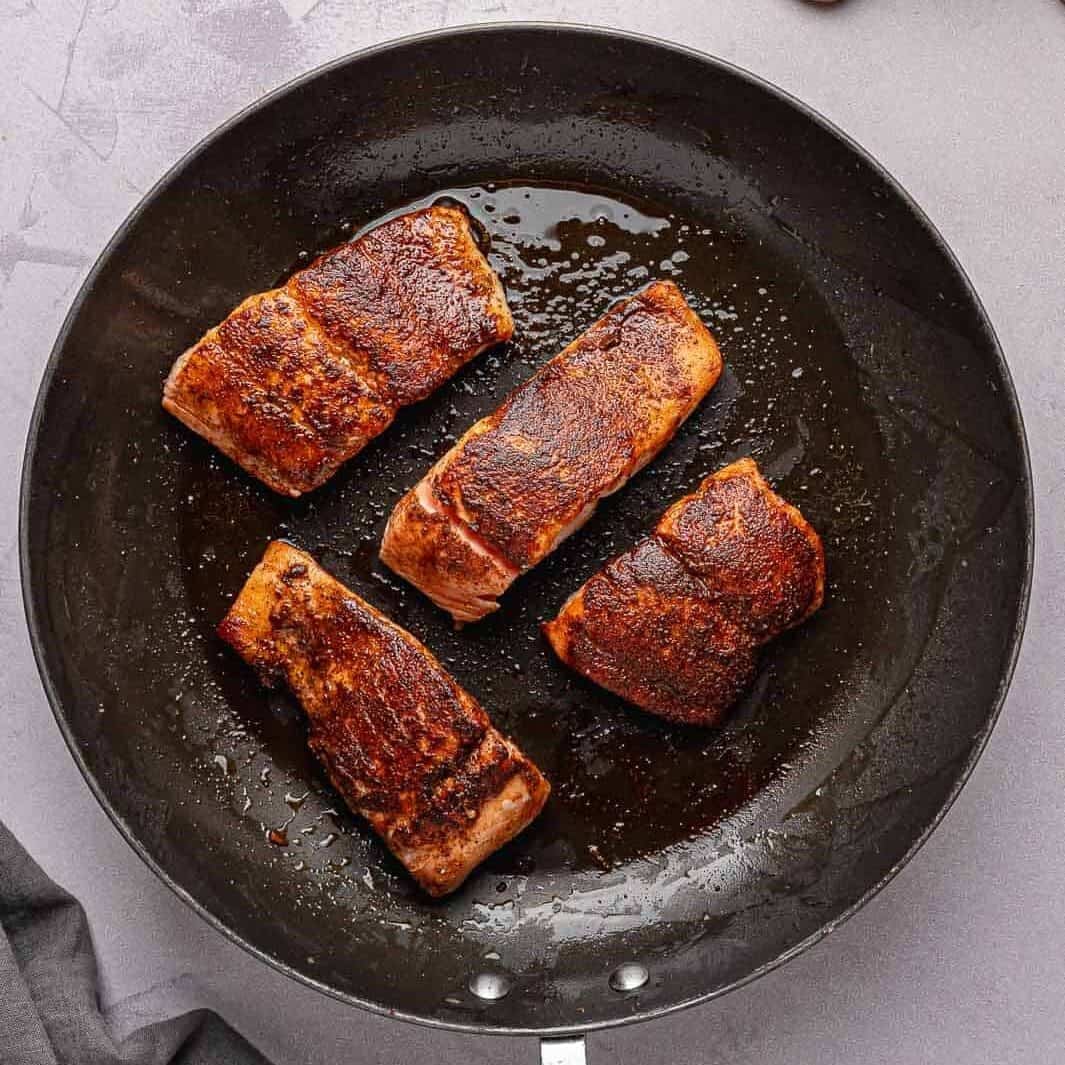 Four of the seasoned salmon fillets are searing in a large, hot non-stick skillet over medium heat. They have developed a crisp, golden-brown crust on one side and are starting to cook through around the edges.