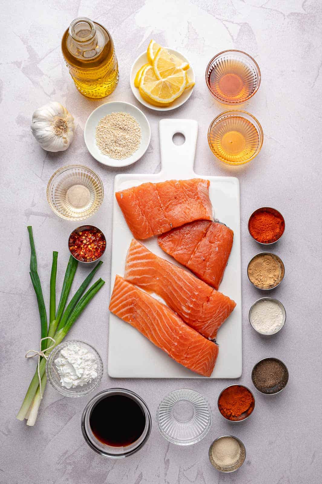 A bright, top-down shot of a clean kitchen counter featuring all the components for the recipe. Four fresh, pink salmon fillets sit on a wooden cutting board next to small glass prep bowls filled with honey, soy sauce, and a colorful pile of dry spices like paprika and cayenne. You can see a few cloves of fresh garlic, a small bottle of sesame oil, and a bunch of vibrant green onions waiting to be chopped. It looks organized, fresh, and ready for cooking.