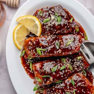 The final, finished Spicy Honey Garlic Salmon in a white serving dish with a thick, caramelized glaze. It is garnished with chopped fresh green onions, toasted sesame seeds, and small lemon wedges.