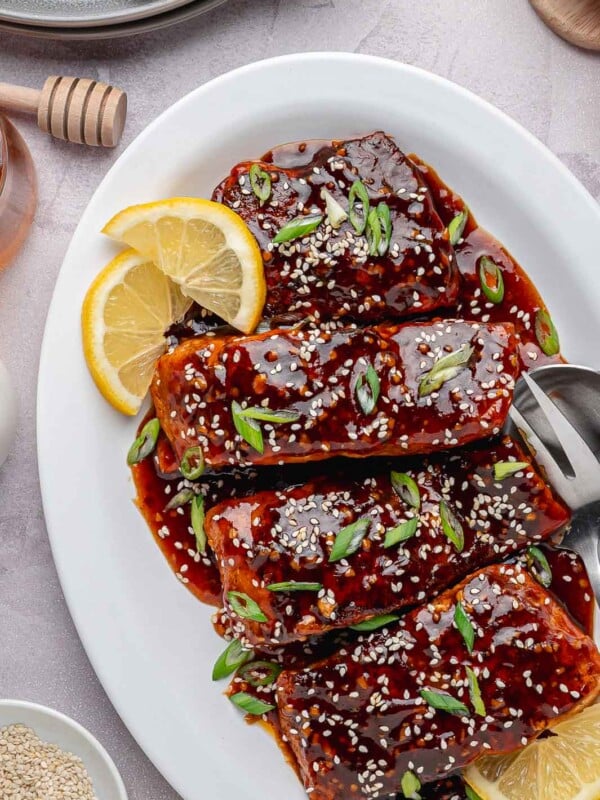 The final, finished Spicy Honey Garlic Salmon in a white serving dish with a thick, caramelized glaze. It is garnished with chopped fresh green onions, toasted sesame seeds, and small lemon wedges.