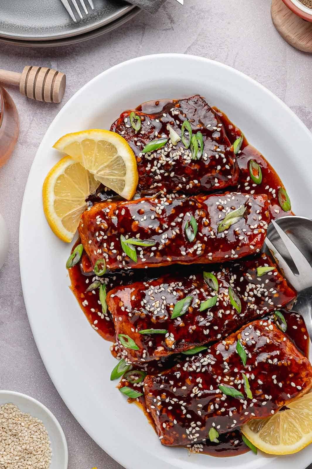 The final, finished Spicy Honey Garlic Salmon in a white serving dish with a thick, caramelized glaze. It is garnished with chopped fresh green onions, toasted sesame seeds, and small lemon wedges.