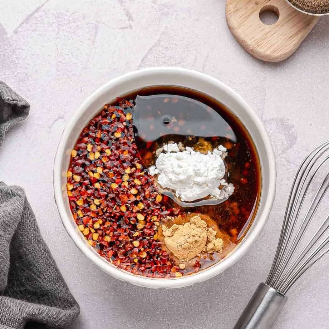 A small bowl on a clean counter where honey, low-sodium soy sauce, rice vinegar, sesame oil, seasonings, minced garlic, corn starch and red chili flakes are being whisked together.