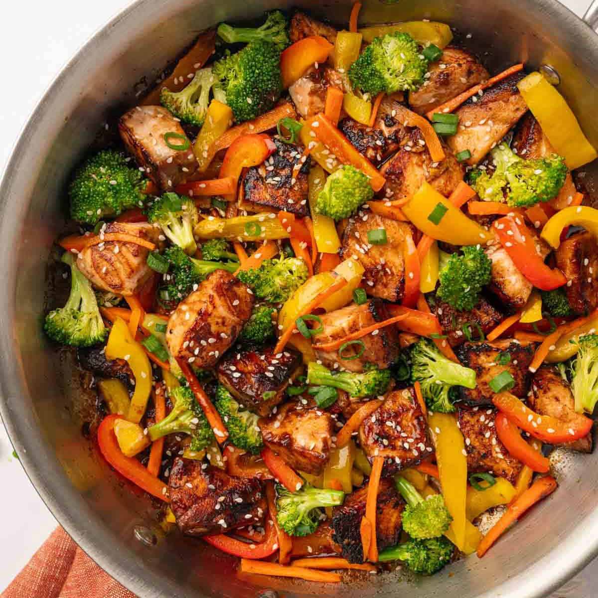 Saut&eacute;ed broccoli, peppers, and carrots being tossed with glazed teriyaki salmon cubes in a large skillet
