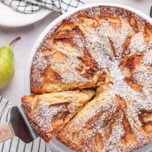 A knife cutting a fresh slice out of a whole round pear cake, showcasing the golden-brown caramelized cinnamon sugar crust.