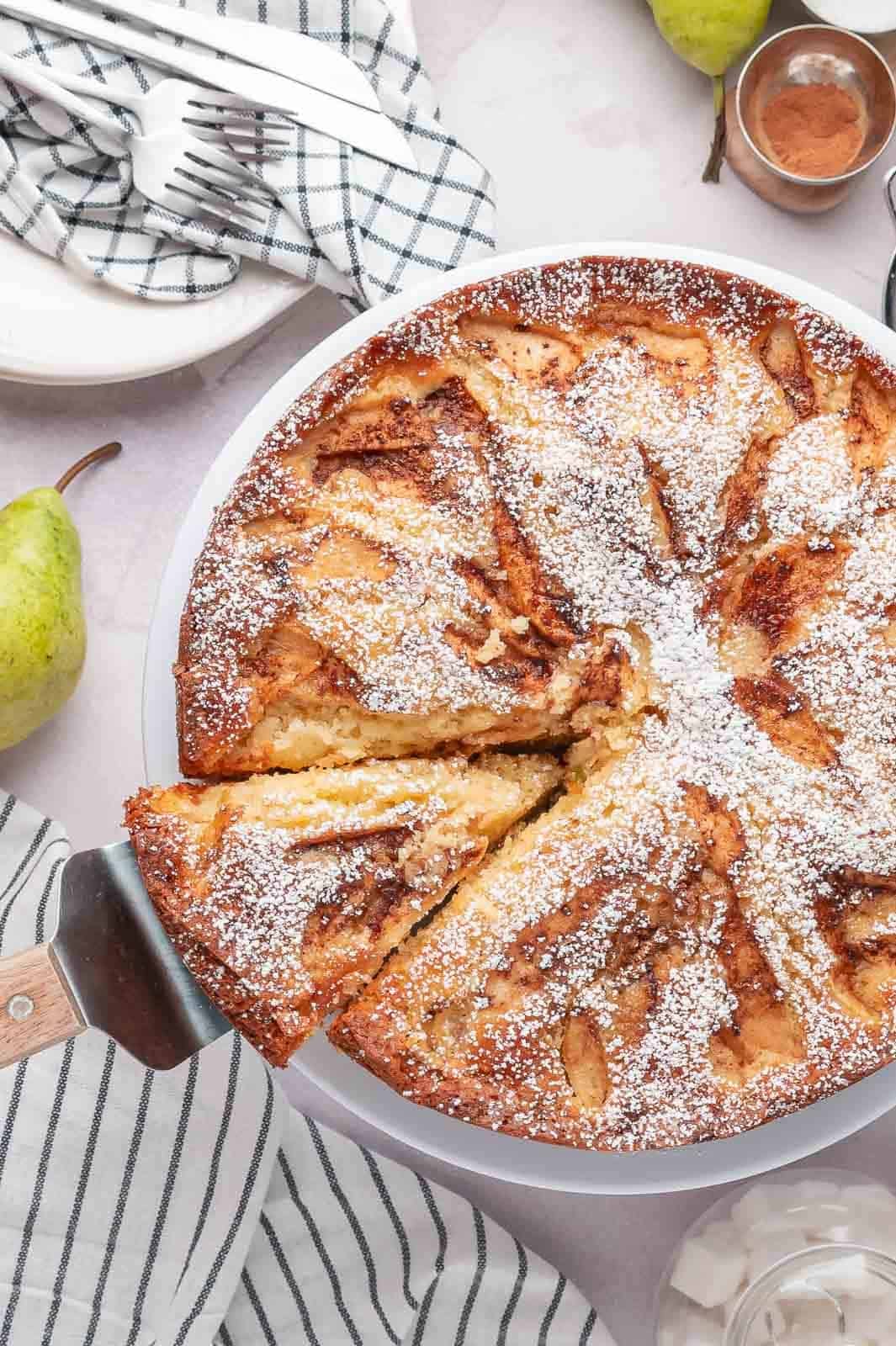 A knife cutting a fresh slice out of a whole round pear cake, showcasing the golden-brown caramelized cinnamon sugar crust.