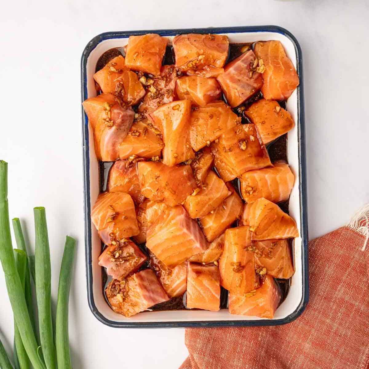 Raw salmon cubes marinating in a shallow glass dish with homemade honey-garlic teriyaki sauce, infusing the fish with flavor before searing.
