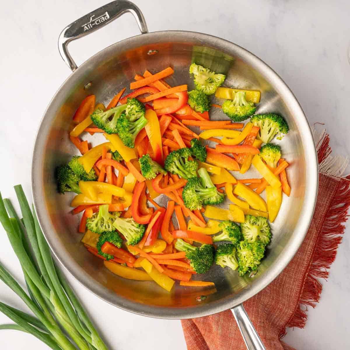 A colorful mix of broccoli florets, sliced red bell peppers, and julienned carrots being tossed in a large skillet over high heat.