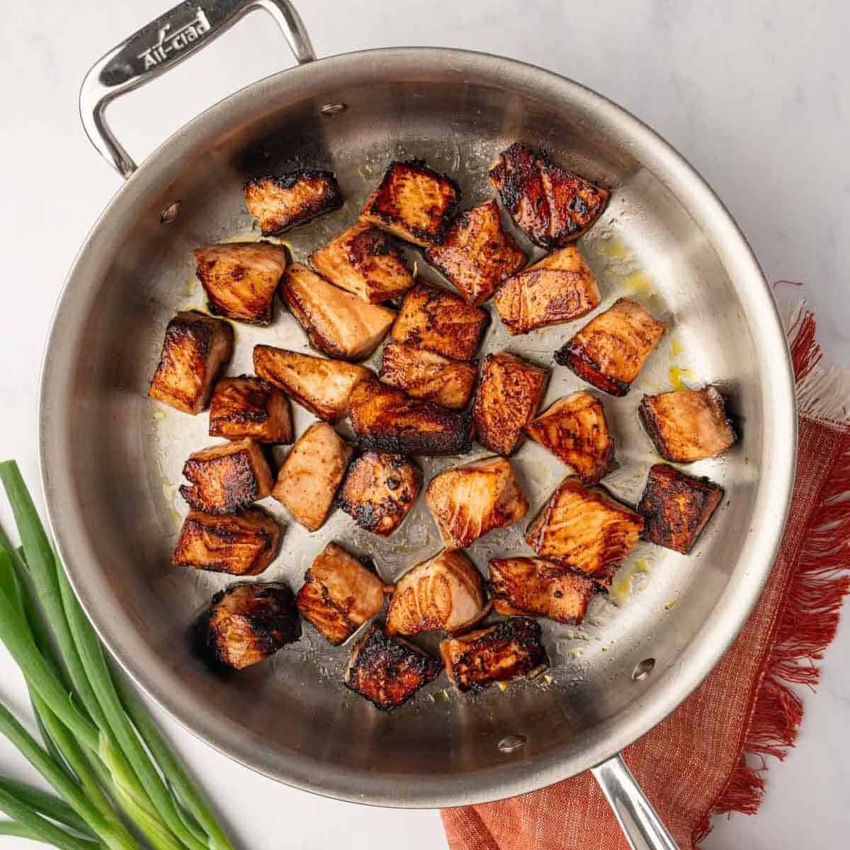 Raw salmon cubes searing in a hot cast-iron skillet with oil, showing the first golden-brown crust forming on the bottom of the fish.