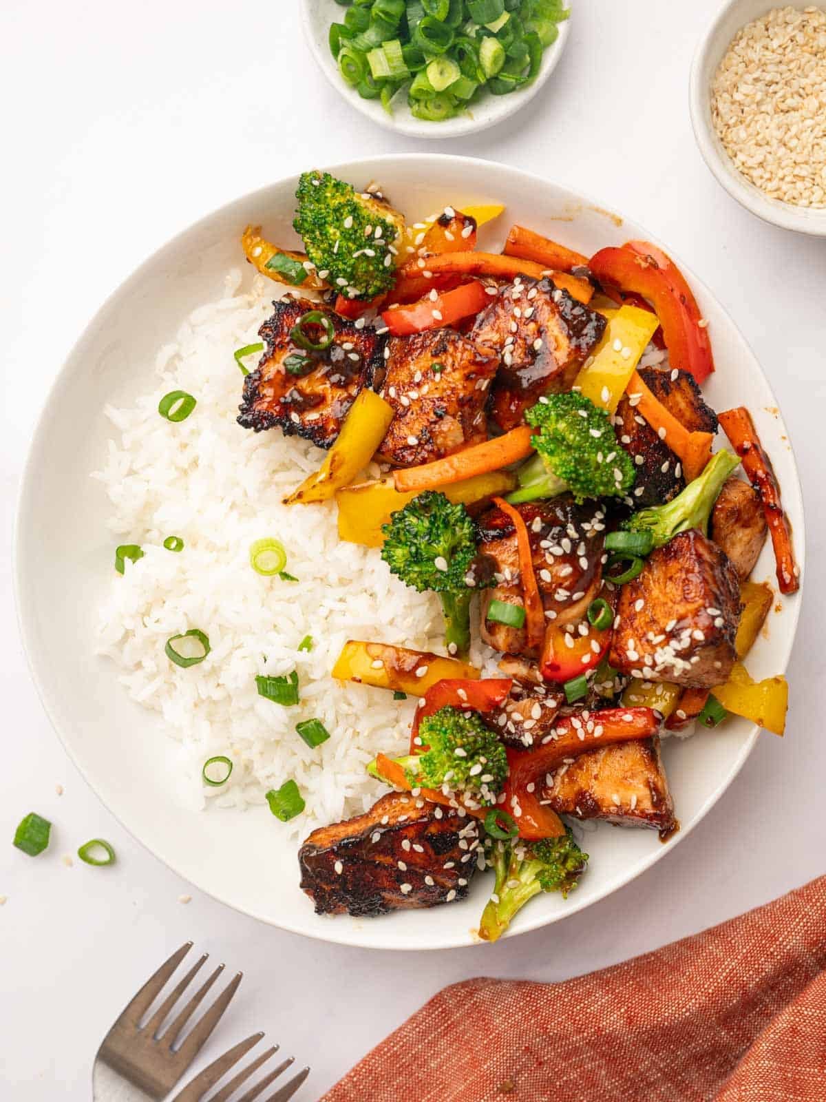 A close-up shot of salmon served with white rice on a plate focusing on glossy, honey-glazed salmon cubes. The teriyaki glaze is thick and coating the fish and vegetables, garnished with white sesame seeds and fresh sliced green onions.