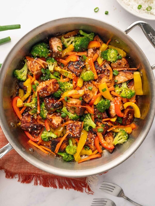 Overhead view of Cookinโ with Mima's Teriyaki Salmon Stir Fry in a large wok, featuring crispy salmon cubes, bright broccoli, and red bell peppers, served over a bed of fluffy jasmine rice.