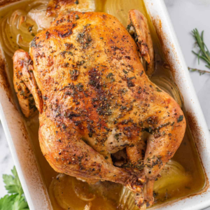 Oven baked whole chicken with golden herb-seasoned skin and onions in the roasting pan.